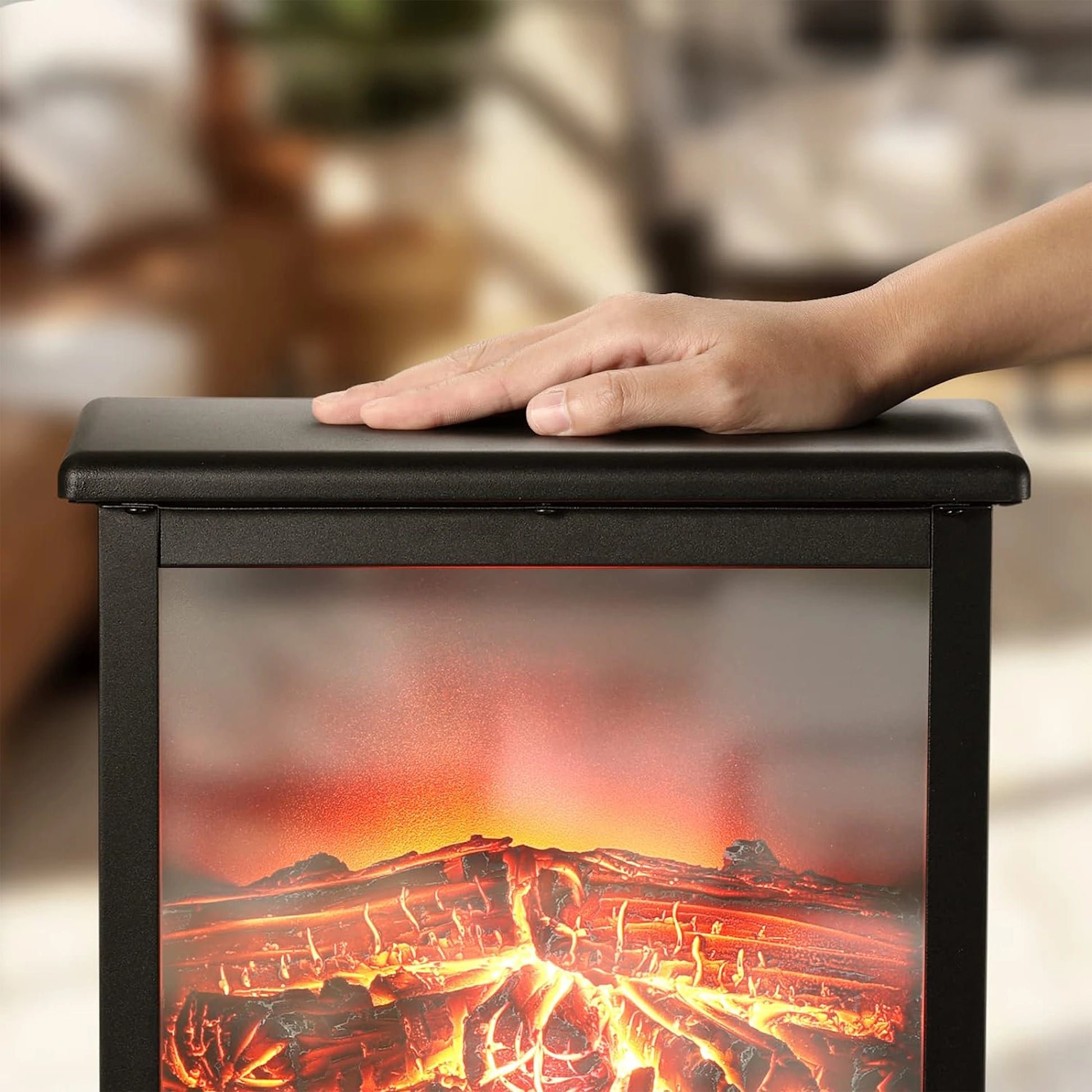 Hand resting on a black box with a fiery design, resembling a fireplace, against a blurred indoor background.